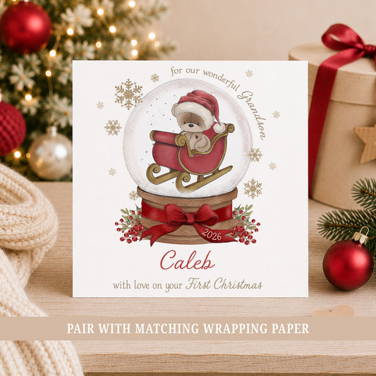Personalised Grandson First Christmas card featuring a teddy bear in a red sleigh inside a Christmas snow globe, with red bow, berries and gold snowflakes. Personalised with a name and relation and also suitable for Son, Nephew or Godson.