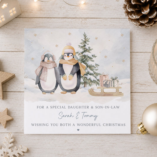Personalised Son and Daughter-in-Law Christmas card featuring a cute penguin couple in a snowy winter scene with Christmas tree, presents and sledge, personalised with both names and available for other couple relations.