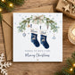A square Christmas greeting card placed on a wooden surface with a brown envelope behind it. The card features an illustrated pair of navy blue stockings labeled 'Mum' and 'Dad,' decorated with green pine branches, gold ornaments, a cream bow, and the message 'Wishing you both a very Merry Christmas.