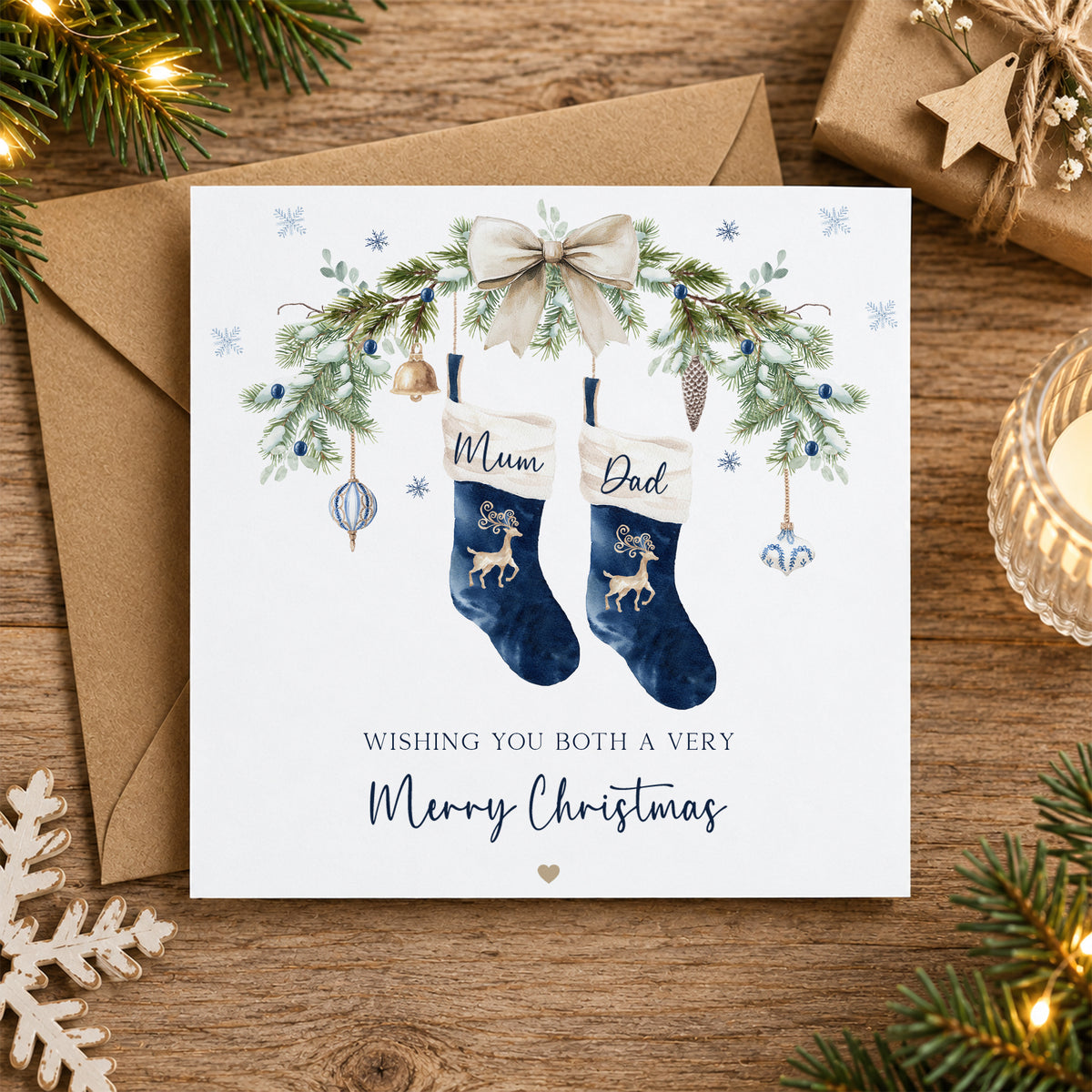A square Christmas greeting card placed on a wooden surface with a brown envelope behind it. The card features an illustrated pair of navy blue stockings labeled 'Mum' and 'Dad,' decorated with green pine branches, gold ornaments, a cream bow, and the message 'Wishing you both a very Merry Christmas.