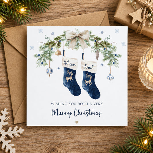 A square Christmas greeting card placed on a wooden surface with a brown envelope behind it. The card features an illustrated pair of navy blue stockings labeled 'Mum' and 'Dad,' decorated with green pine branches, gold ornaments, a cream bow, and the message 'Wishing you both a very Merry Christmas.