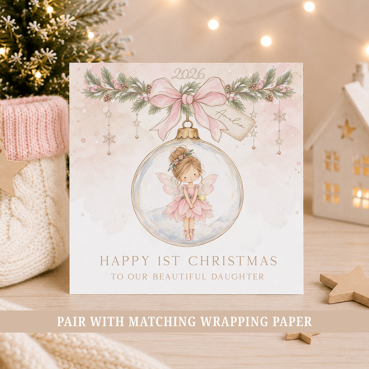 A square Christmas greeting card displayed on a wooden surface with holiday decor in the background. The card features a watercolor illustration of a fairy inside a pink ornament, with a pink bow and green garland at the top.