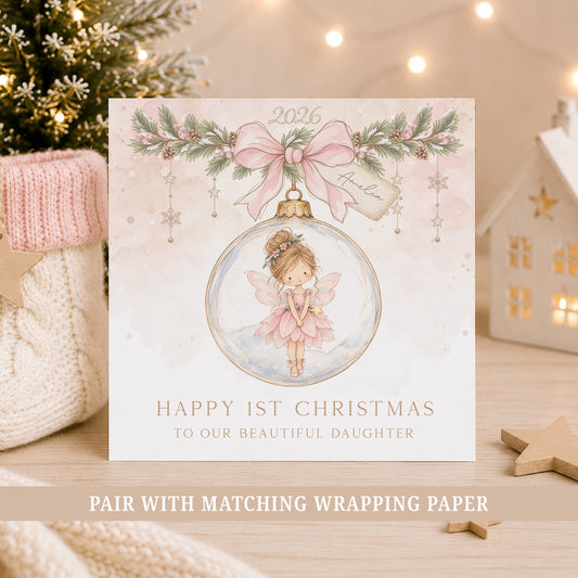 A square Christmas greeting card displayed on a wooden surface with holiday decor in the background. The card features a watercolor illustration of a fairy inside a pink ornament, with a pink bow and green garland at the top.