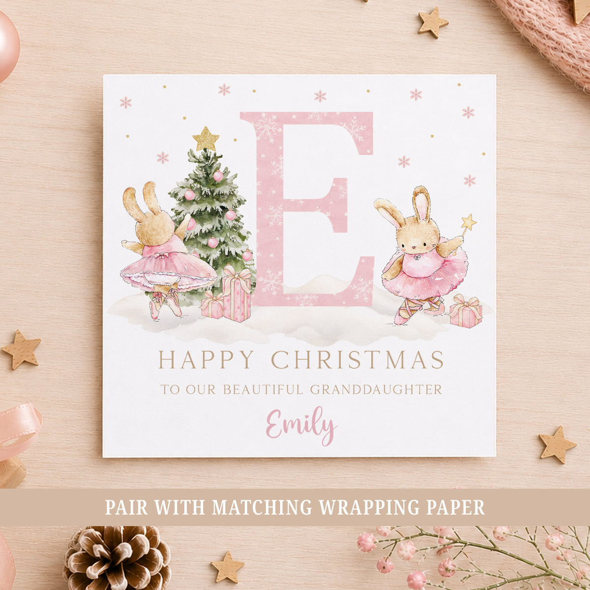 Personalised ballerina bunny Christmas card for girls, featuring two watercolour bunnies wearing pink tutus and ballet shoes beside a decorated Christmas tree with pink baubles, a gold star and wrapped presents. A large pink initial with a snowflake pattern is displayed in the centre, with pink snowflakes and gold details throughout. Personalised with the recipient's name and relation, shown with the letter E for Granddaughter Emily.