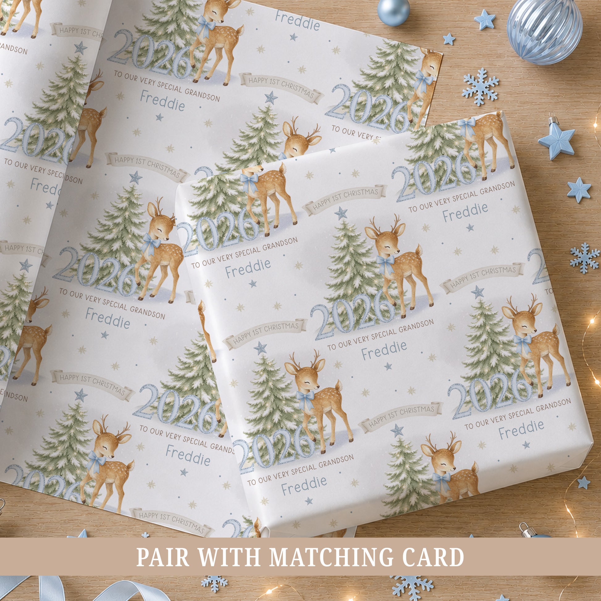 Personalised baby boy First Christmas wrapping paper featuring a reindeer with blue bow, snowy Christmas tree, blue 2026 numbers, stars and snowflakes.