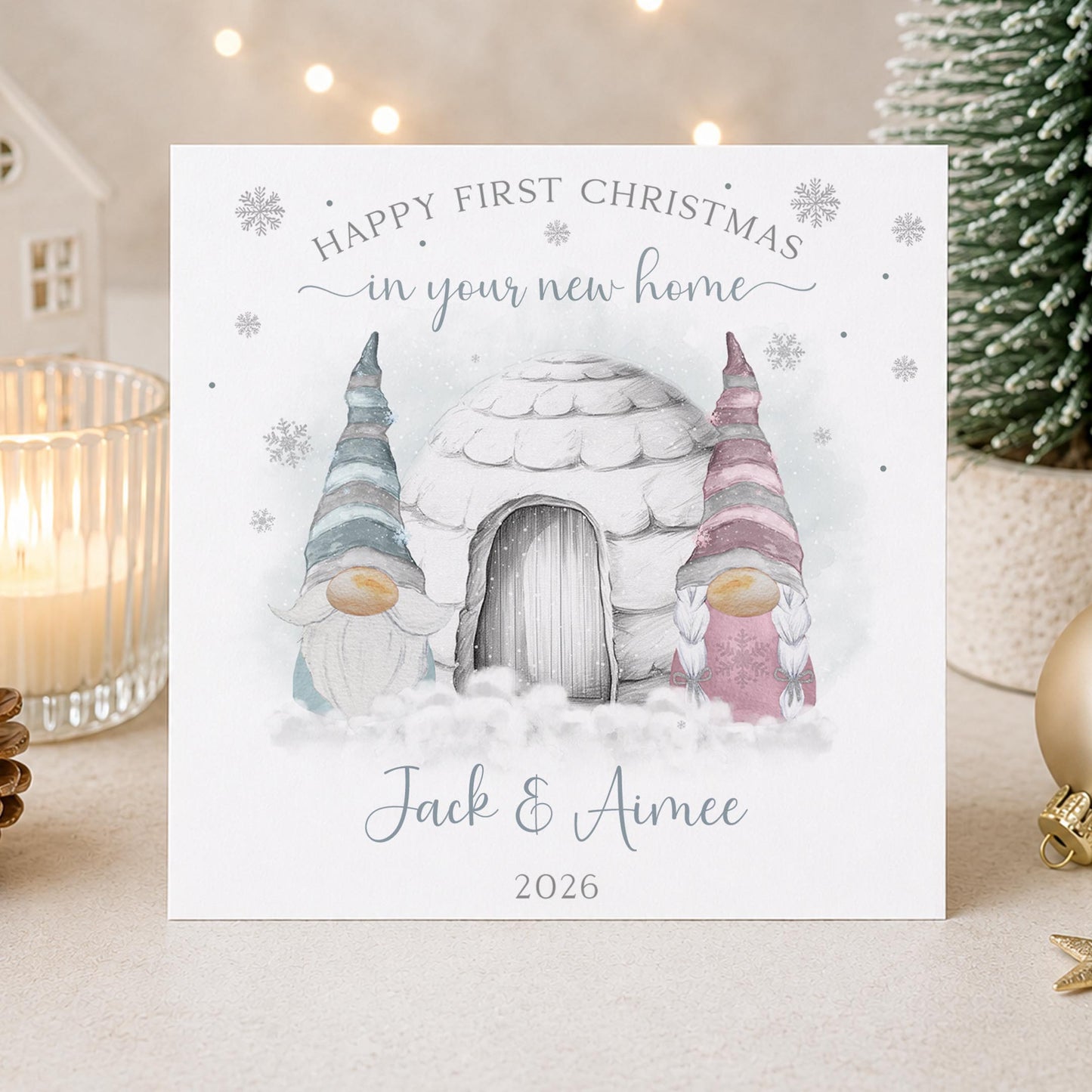 A personalised Christmas greeting card displayed on a tabletop with holiday decor in the background. The card features an illustration of a snowy cottage and two watercolor-style gnomes, with the text 'HAPPY FIRST CHRISTMAS in your new home' and the plus the year '2026'.