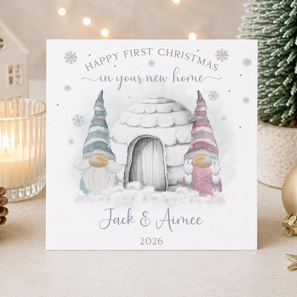 A personalised Christmas greeting card displayed on a tabletop with holiday decor in the background. The card features an illustration of a snowy cottage and two watercolor-style gnomes, with the text 'HAPPY FIRST CHRISTMAS in your new home' and the plus the year '2026'.