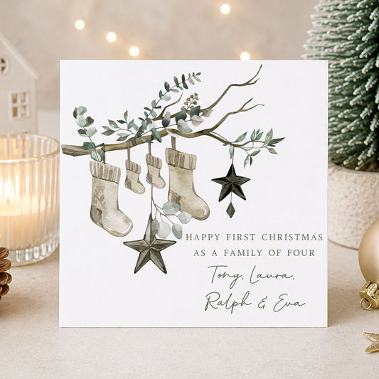 Personalised first Christmas as a family of four card featuring four festive stockings hanging from a eucalyptus branch with black star decorations and personalised family names.