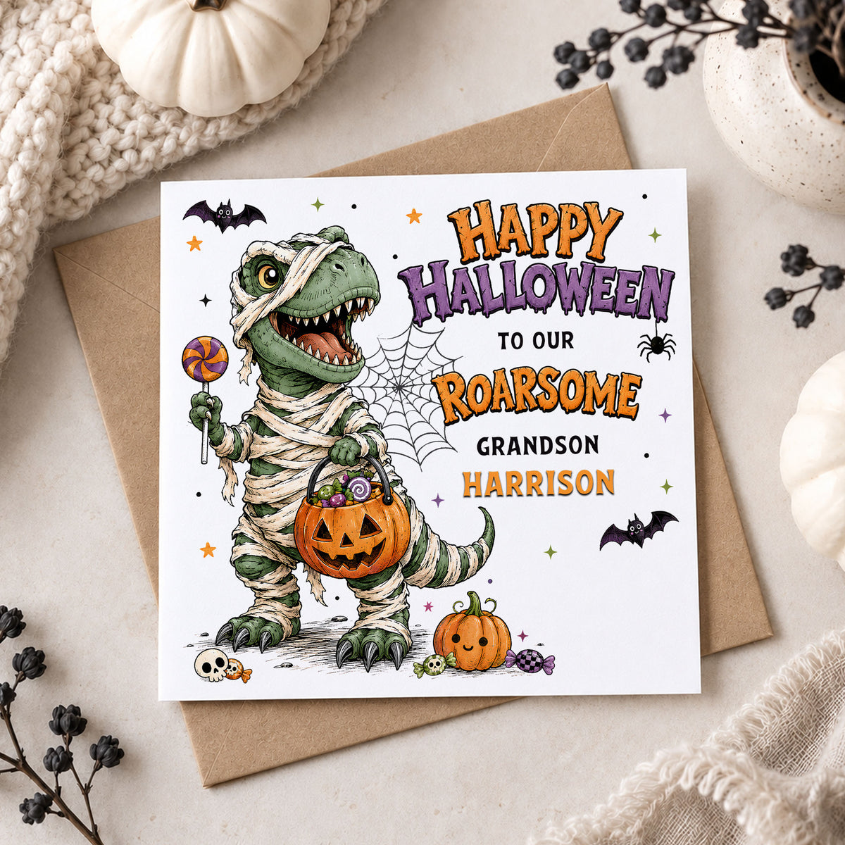 Personalised Grandson Halloween card featuring a green dinosaur dressed as a mummy, holding a pumpkin trick or treat bucket and lollipop, with bats, spider web, pumpkins and sweets. Personalised with his name and available for Grandson, Son, Nephew, Godson, Little Boy and other relations.