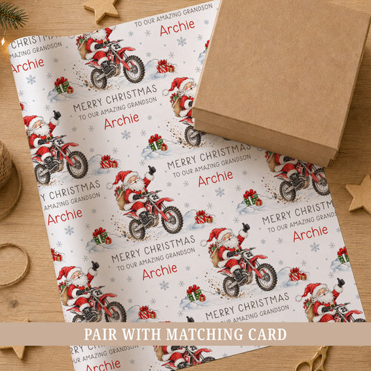 Personalised Christmas wrapping paper featuring Santa riding a red motocross dirt bike through the snow with festive presents and snowflakes, personalised with a name and choice of relation.